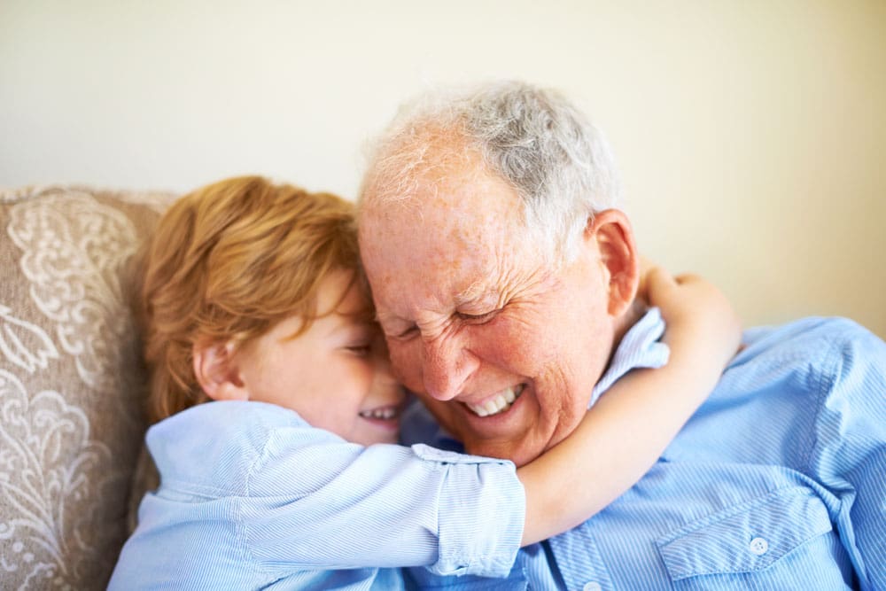 Young boy hugs older memory care resident while they both smile with their eyes closed 